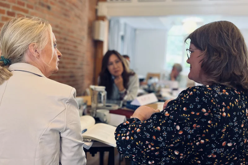 To kvinder sidder og diskuterer ved et bord i et mødelokale, mens en tredje kvinde lytter i baggrunden. Der er bøger og papirer spredt på bordet, hvilket skaber en atmosfære af faglig dialog og samarbejde.