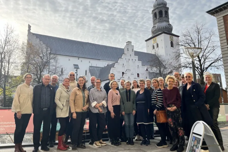 Berigende dage i Aalborg "Gruppefoto af en større forsamling mennesker foran Budolfi Kirke i Aalborg på en solrig dag. Personerne smiler og poserer foran den historiske bygning, der har en markant klokketårn og hvidkalkede mure."