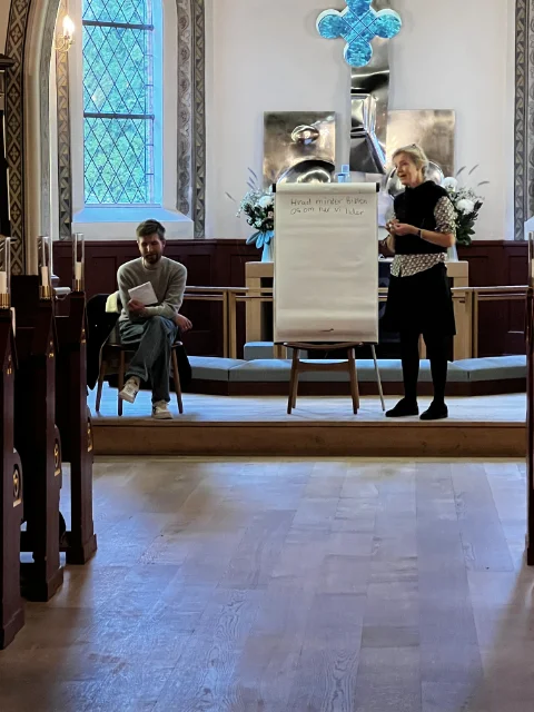 En præst og en person sidder foran en flipover i en kirke. Der er smukt dekoreret med blomster og et kors i baggrunden. Flipoveren bruges til en præsentation eller undervisning for de fremmødte.