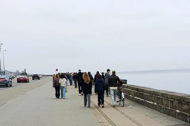 Er bibelsk sjælesorg også for konfirmander En gruppe konfirmander går på en promenade ved vandet. Det ser ud til at være en social aktivitet eller en del af en ceremoni, i et roligt og gråt vejr.