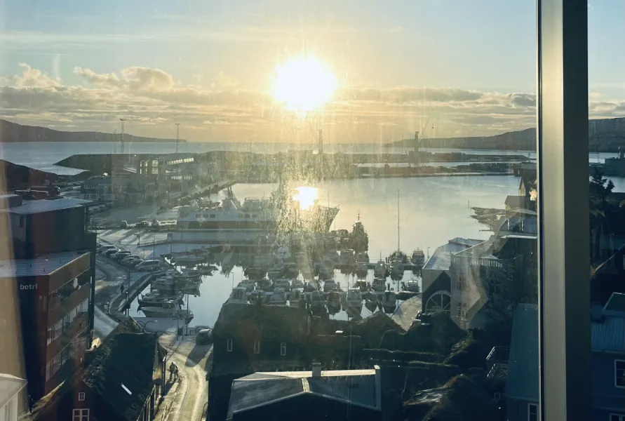 Udsigt over en havn med både og bygninger, set gennem et vindue ved solnedgang, hvor solen skinner over vandet.