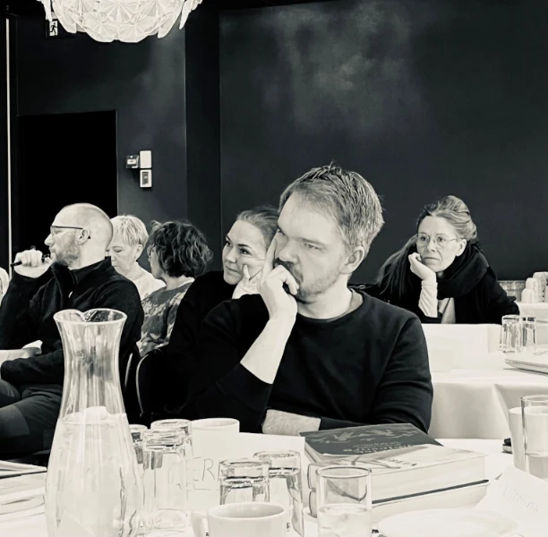 Sort-hvidt billede af personer til en konference eller workshop, hvor en mand i forgrunden ser tænksom ud.