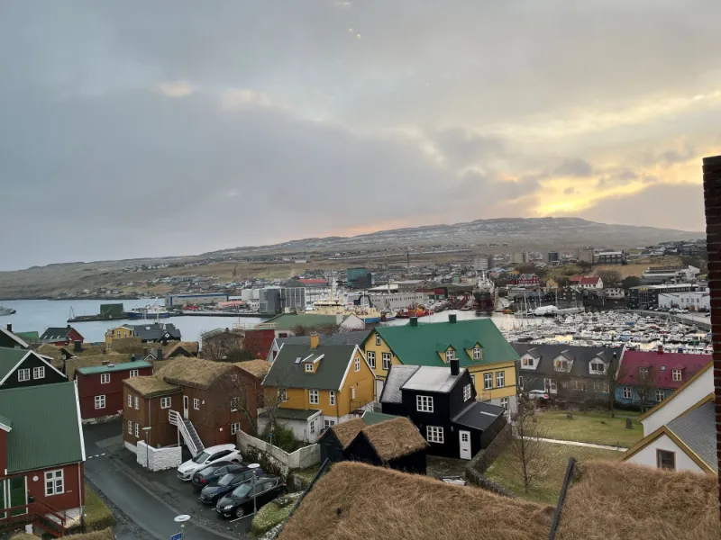Udsigt over en færøsk by med farverige huse, havn og bjerge i baggrunden ved solnedgang.