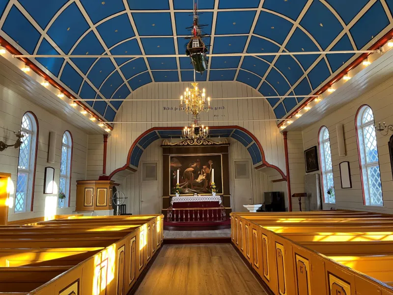 Interiør af en traditionel kirke med blå loftsdekoration, træbænke og et maleri ved alteret.