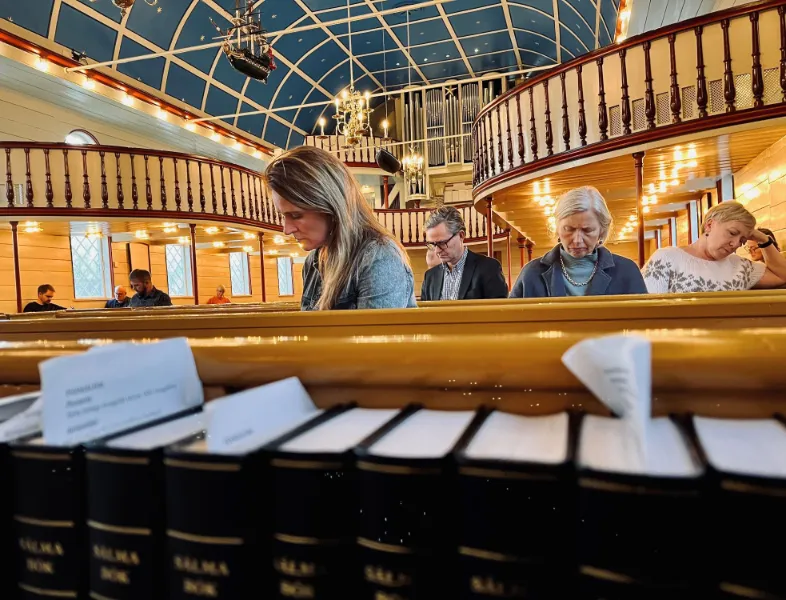 Indre af en kirke med folk, der sidder på bænke, omgivet af salmebøger og et dekoreret loft.