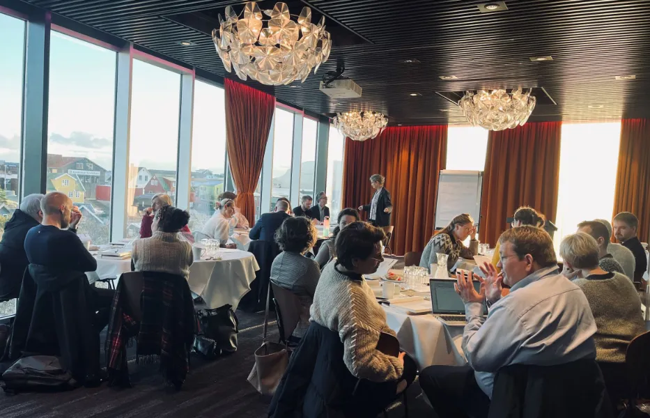 Konference eller workshop i en moderne mødesal med store vinduer, hvor flere personer sidder ved runde borde og deltager i diskussioner.