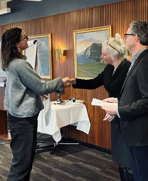 Kvinde giver håndtryk og modtager et certifikat fra to personer i et træbeklædt rum med malerier på væggene.
