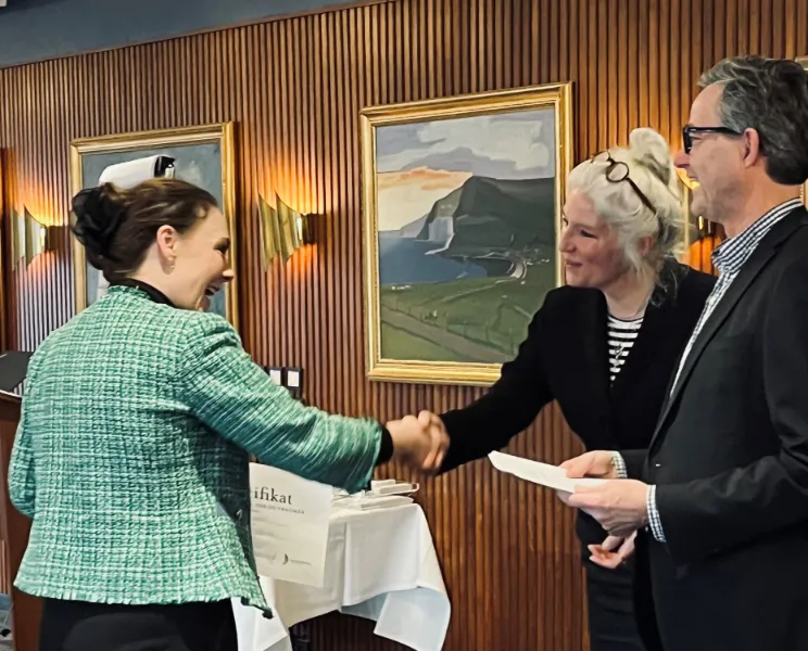 Kvinde giver håndtryk og modtager et certifikat fra to personer i et træbeklædt rum.