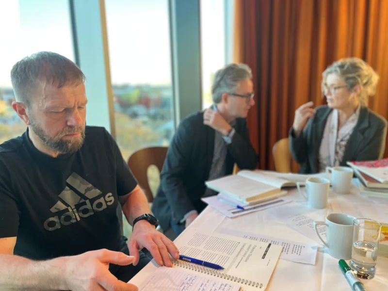 Mand i sort Adidas-T-shirt skriver i en notesbog, mens to andre personer i jakkesæt diskuterer i baggrunden ved et bord med bøger og kaffekopper.