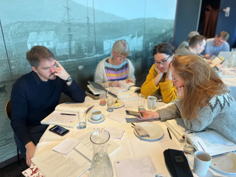 Gruppe af fire personer sidder ved et bord med bøger, noter og kaffekopper, mens de arbejder koncentreret og diskuterer i en konference- eller workshop-setting.