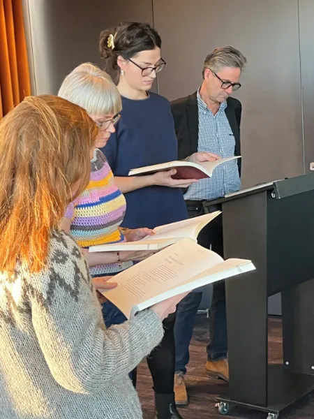 Fire personer står ved en talerpult og læser højt fra bøger i en formel indendørs setting.