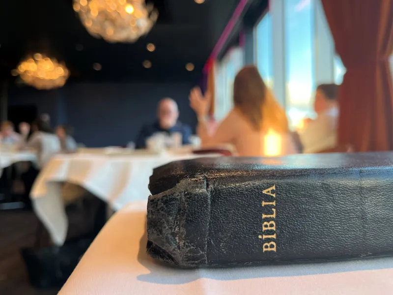Slidt bibel med ordet "BIBLIA" præget på ryggen ligger på et bord, med en gruppe mennesker i baggrunden, der deltager i en diskussion i et konferencelokale.