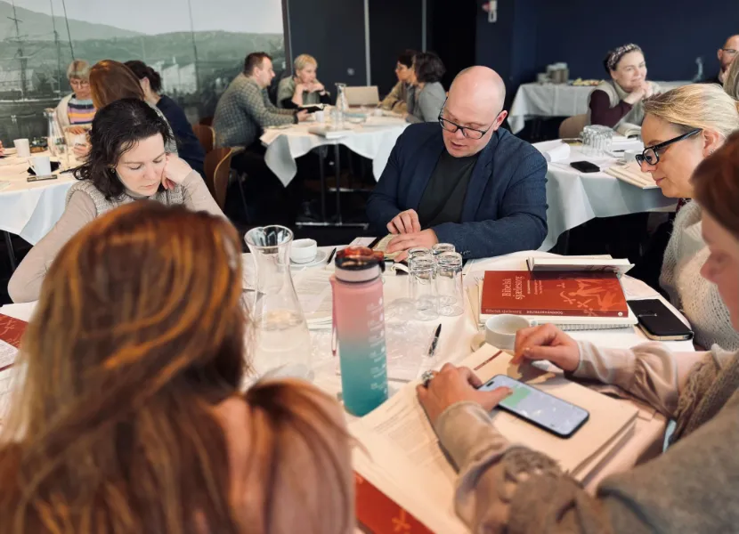 Gruppe af personer sidder ved et rundt bord omgivet af bøger, papirer og vandglas, mens de arbejder og diskuterer i en konference- eller workshop-setting.