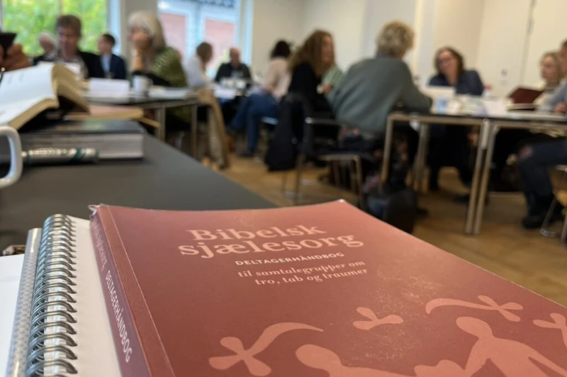 Bog med titlen "Bibelsk sjælesorg" ligger på et bord i forgrunden, mens en gruppe mennesker sidder samlet til en diskussion i baggrunden.
