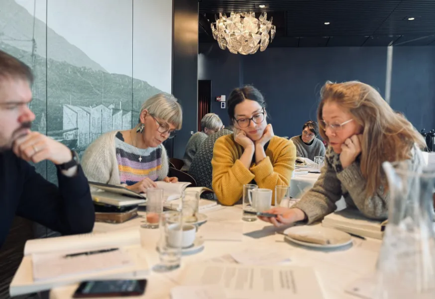 En gruppe mennesker sidder omkring et bord i en moderne restaurant, engageret i en diskussion med dokumenter og kopper på bordet.