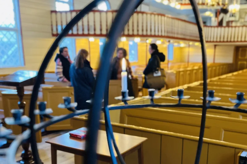 Indvendigt billede af en traditionel trækirke med gule bænke og en sort smedejernskandelaber i forgrunden. En gruppe mennesker står og samtaler i baggrunden.