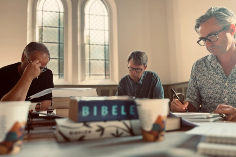 Tre mænd sidder koncentreret og læser i bibler ved et bord i et lyst kirkerum med buede vinduer i baggrunden.