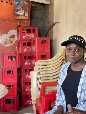 Ung kvinde sidder ved stablede Coca-Cola-kasser og plaststole i et farverigt hjørne med vægmaleri og religiøst billede.