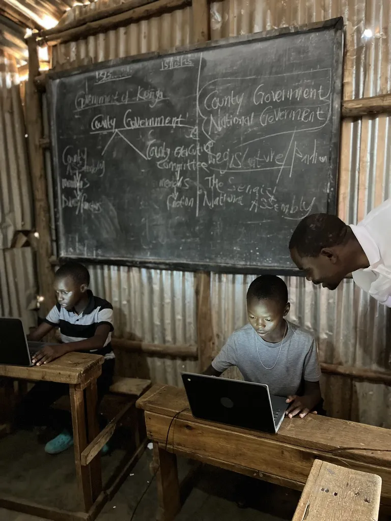 To drenge arbejder ved bærbare computere i et klasselokale med træborde og tavle i en simpel bygning, mens en lærer assisterer.