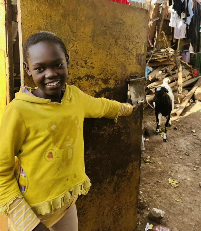 Pige i gul trøje holder i en metaldør med en sort og hvid ged i baggrunden i et gårdmiljø med træstabler og vasketøj.