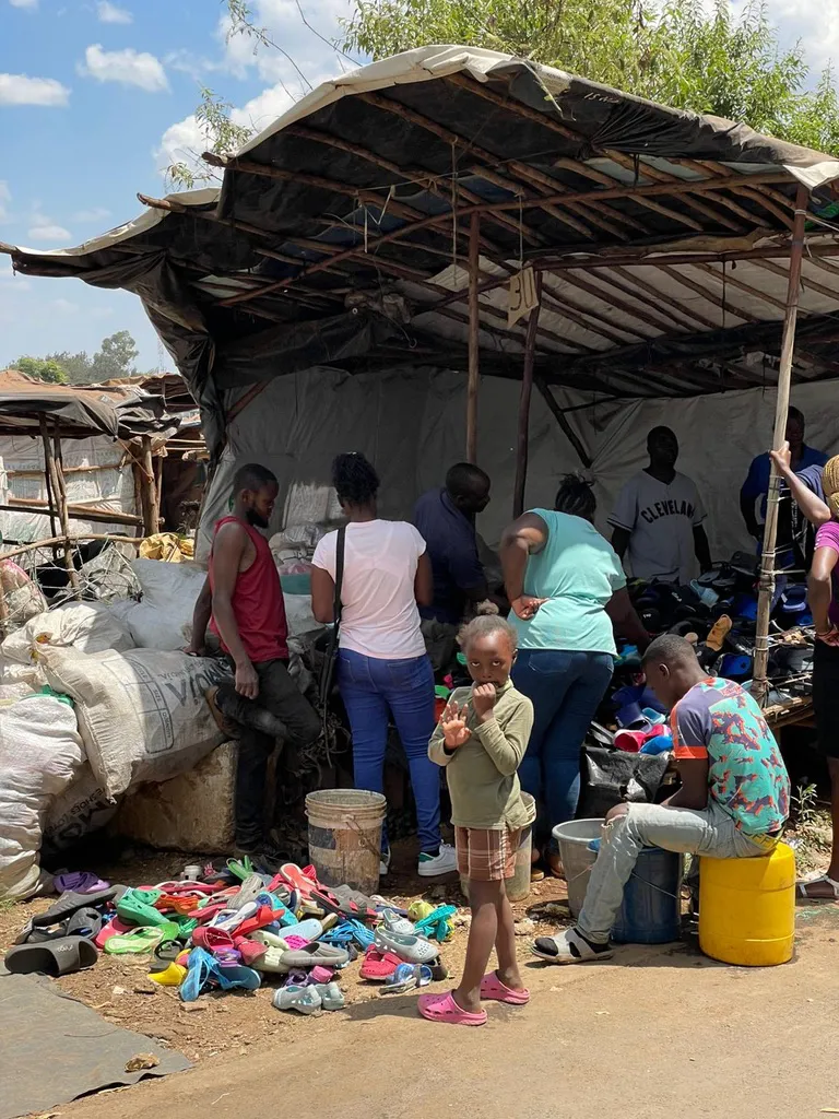 Gruppe mennesker handler ved en gadebod med brugt fodtøj, mens en lille pige står foran og ser mod kameraet.