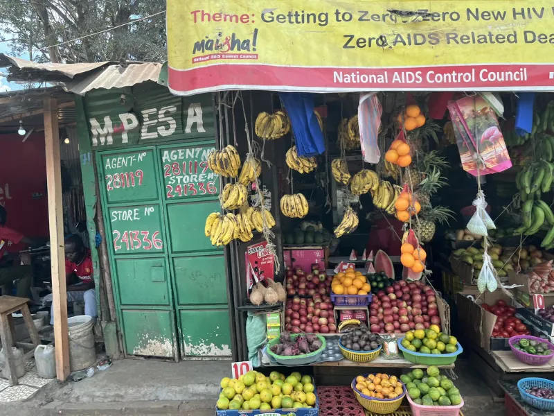 Frugt- og grøntbod med bananer, appelsiner og avocadoer foran en M-PESA kiosk, med banner fra National AIDS Control Council.