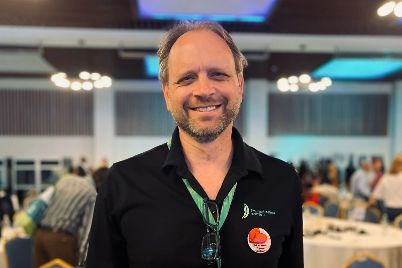 Philip G. Monroe, der smiler til kameraet i en stor sal. Han er iført en sort poloshirt med "Trauma Healing Institute" logoet, en grøn snor om halsen og briller, der hænger i kraven. I baggrunden ses et sløret lokale med mennesker og dækkede borde.