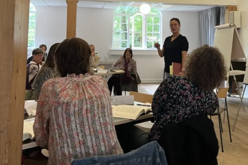 Kursus i lyst lokale med voksne deltagere ved borde, mens en kvinde underviser foran en flipover.