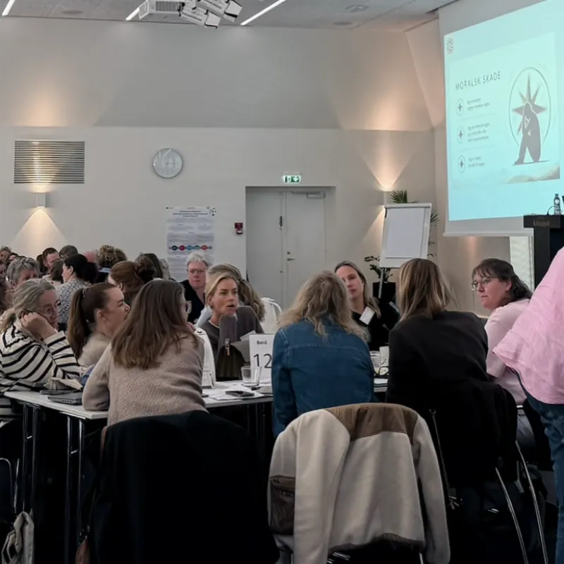 Deltagere arbejder i grupper ved borde under et seminar om moralsk skade, mens en præsentation vises på lærredet.
